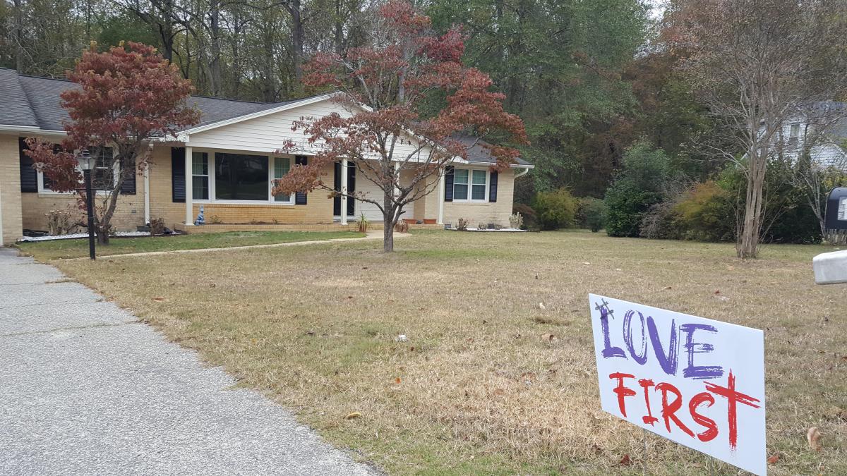 Love First sign in a customers yard with a beige house in the background